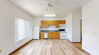 This efficiently designed single-floor unit offers a well-utilized open-concept layout that combines the kitchen and living areas into one cohesive space. Light wood laminate flooring runs throughout, lending warmth and visual continuity to the interior.

The kitchen is equipped with warm-toned wood cabinetry—both upper and lower—providing generous storage. Dark countertops offer a subtle contrast, while white appliances, including a refrigerator, electric range, and dishwasher, contribute to a clean, functional aesthetic. A ceiling fan overhead adds comfort and airflow, and multiple windows allow natural light to brighten the space while framing views of the surrounding neighborhood.

The living area is open and airy with crisp white walls, high ceilings that enhance the sense of volume, and large windows fitted with vertical blinds that allow for adjustable light control. The layout offers a versatile canvas for a variety of furniture arrangements.

The bathroom features light wood flooring consistent with the rest of the unit, a wall-mounted vanity with a blue countertop, a large mirror, and a full bathtub with a shower, complemented by a grab bar for added accessibility. A wooden cabinet provides additional storage. Overall, the unit presents a clean, functional, and well-lit living environment.