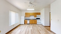 This efficiently designed single-floor unit offers a well-utilized open-concept layout that combines the kitchen and living areas into one cohesive space. Light wood laminate flooring runs throughout, lending warmth and visual continuity to the interior.

The kitchen is equipped with warm-toned wood cabinetry—both upper and lower—providing generous storage. Dark countertops offer a subtle contrast, while white appliances, including a refrigerator, electric range, and dishwasher, contribute to a clean, functional aesthetic. A ceiling fan overhead adds comfort and airflow, and multiple windows allow natural light to brighten the space while framing views of the surrounding neighborhood.

The living area is open and airy with crisp white walls, high ceilings that enhance the sense of volume, and large windows fitted with vertical blinds that allow for adjustable light control. The layout offers a versatile canvas for a variety of furniture arrangements.

The bathroom features light wood flooring consistent with the rest of the unit, a wall-mounted vanity with a blue countertop, a large mirror, and a full bathtub with a shower, complemented by a grab bar for added accessibility. A wooden cabinet provides additional storage. Overall, the unit presents a clean, functional, and well-lit living environment.