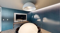 This intimate conference room is a thoughtfully designed meeting space that balances creative aesthetics with functional purpose. The room's most distinctive feature is its deep teal accent walls adorned with panel molding and an artistic cloud mural, lending the space a sophisticated, sky-inspired atmosphere. A large wall-mounted flat-screen display is paired with a stylish wooden credenza, making the room well-equipped for presentations and collaborative meetings. At the center of the room sits a sleek oval white conference table surrounded by plush dark upholstered chairs, providing comfortable seating for small group gatherings. Overhead, a dramatic oversized dome pendant light with a silver metallic interior serves as a bold focal point, complemented by subtle cove lighting that traces the perimeter of the ceiling, adding a warm, layered glow. The floor is finished with a textured, patterned area rug that anchors the space and adds visual interest. A floor-to-ceiling frosted glass sliding partition wall connects the room to the adjacent open office area, allowing for visual privacy while maintaining a sense of openness and transparency. The overall ambiance is polished and modern, making it an ideal setting for focused, professional collaboration.