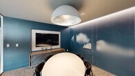 This intimate conference room is a thoughtfully designed meeting space that balances creative aesthetics with functional purpose. The room's most distinctive feature is its deep teal accent walls adorned with panel molding and an artistic cloud mural, lending the space a sophisticated, sky-inspired atmosphere. A large wall-mounted flat-screen display is paired with a stylish wooden credenza, making the room well-equipped for presentations and collaborative meetings. At the center of the room sits a sleek oval white conference table surrounded by plush dark upholstered chairs, providing comfortable seating for small group gatherings. Overhead, a dramatic oversized dome pendant light with a silver metallic interior serves as a bold focal point, complemented by subtle cove lighting that traces the perimeter of the ceiling, adding a warm, layered glow. The floor is finished with a textured, patterned area rug that anchors the space and adds visual interest. A floor-to-ceiling frosted glass sliding partition wall connects the room to the adjacent open office area, allowing for visual privacy while maintaining a sense of openness and transparency. The overall ambiance is polished and modern, making it an ideal setting for focused, professional collaboration.