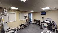 This fitness room offers a practical and functional workout environment designed to accommodate a variety of exercise needs. The space features a drop ceiling with recessed panel lighting that provides consistent, even illumination throughout. The flooring is finished with durable, dark rubber gym matting, ideal for supporting heavy equipment and high-impact activity. A elliptical cross-trainer takes a prominent position on the floor, complemented by additional cardio equipment nearby. A multi-tiered dumbbell rack lines one wall, stocked with a range of free weights to support strength training at various levels. A wall-mounted flat-screen television is positioned for easy viewing during workouts. The neutral beige walls and frosted windows contribute to a clean, focused atmosphere, while a glass-paneled door provides access to the adjacent corridor, allowing for natural light to filter in. A hand sanitizer dispenser is conveniently mounted near the entrance, reflecting attention to hygiene. Overall, the space is efficiently laid out to maximize usability within its footprint, making it a well-considered amenity for fitness-minded users.