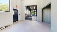 Located on Bond Street in Bridgeport, Connecticut, this single-floor commercial space offers a generously proportioned open-plan layout designed for multi-functional use. The space showcases an industrial-modern aesthetic, defined by soaring exposed ceilings with visible structural grid work, pendant track lighting, and a striking teal-painted ceiling that adds bold visual character throughout the main lounge area.

The primary lounge zone is furnished with clusters of plush velvet sofas and seating arrangements, set against deep green-painted masonry walls accented with vertical wood panel detailing. Large, vertically oriented windows run along multiple walls, flooding the interior with abundant natural light and providing views of the surrounding greenery and streetscape.

A dedicated fitness area is accessible through a wide open passage, equipped with treadmills and cardio equipment, with full-height mirrors, bright overhead lighting, and a lush green moss wall feature adding a fresh, energetic atmosphere. A quieter nook area features minimalist wood furniture suited for small meetings or breakout sessions.

The polished light-toned large-format tile flooring extends uniformly throughout, unifying the various zones. Painted concrete block walls in the utility areas and visible mechanical infrastructure reinforce the raw, industrial character of the space. With its open layout and versatile zones, this space is well-suited for community, wellness, or creative commercial use.
