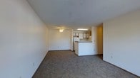 Located at 450 Kristina Dr in Bellefontaine, Ohio, this single-floor unit offers a practical and comfortable layout designed for everyday living. The open living area is finished with wall-to-wall carpet flooring, providing a warm and inviting atmosphere. A ceiling fan with a light fixture serves as the central overhead lighting for the space, contributing to a simple and functional design.

The living area flows seamlessly into the kitchen, which is defined by a built-in peninsula that creates a natural division between the two spaces while maintaining an open feel. The kitchen is equipped with upper cabinetry featuring dark wood trim and a full-size refrigerator, offering solid storage and utility. Recessed fluorescent lighting above the kitchen work area ensures ample illumination for meal preparation.

The interior palette is kept neutral throughout, with white walls that reflect light and create a clean, uncluttered look. The overall layout is straightforward and efficient, making good use of the available floor space. The single-level design adds to the accessibility and ease of navigation throughout the unit, making it a practical option for a variety of occupants.