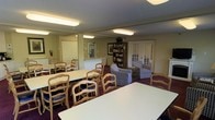 This single-floor community space is thoughtfully designed to serve as both a dining and social gathering area. The room is generously sized, accommodating multiple rectangular white dining tables surrounded by classic wood-framed chairs with blue upholstered cushions, providing ample seating for group activities or shared meals. Overhead, flush-mounted fluorescent lighting ensures the space is well-lit throughout.

A cozy lounge corner is set apart from the dining area, furnished with patterned armchairs arranged invitingly around a low coffee table, creating a comfortable nook for relaxed conversation. A tall wooden bookshelf stands nearby, filled with books and adding a homey, lived-in character to the space.

The walls are finished in a warm neutral tone, complemented by dark burgundy carpeting that grounds the room with a sense of warmth. Framed artwork decorates the walls, contributing to the welcoming ambiance. Multiple doorways — including a classic paneled door and a set of French doors allowing a glimpse of natural light from outside — connect the space to adjacent areas, enhancing its accessibility and openness. Overall, the space strikes a balance between functional communal use and comfortable, informal living.