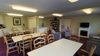 This single-floor community space is thoughtfully designed to serve as both a dining and social gathering area. The room is generously sized, accommodating multiple rectangular white dining tables surrounded by classic wood-framed chairs with blue upholstered cushions, providing ample seating for group activities or shared meals. Overhead, flush-mounted fluorescent lighting ensures the space is well-lit throughout.

A cozy lounge corner is set apart from the dining area, furnished with patterned armchairs arranged invitingly around a low coffee table, creating a comfortable nook for relaxed conversation. A tall wooden bookshelf stands nearby, filled with books and adding a homey, lived-in character to the space.

The walls are finished in a warm neutral tone, complemented by dark burgundy carpeting that grounds the room with a sense of warmth. Framed artwork decorates the walls, contributing to the welcoming ambiance. Multiple doorways — including a classic paneled door and a set of French doors allowing a glimpse of natural light from outside — connect the space to adjacent areas, enhancing its accessibility and openness. Overall, the space strikes a balance between functional communal use and comfortable, informal living.