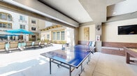 Located in Escondido, California, this amenity-rich community space offers a seamless blend of indoor and outdoor recreation. The interior lounge area features a tray ceiling with recessed lighting and a modern ceiling fan, warm wood-paneled accent walls, and a built-in wet bar complete with a beverage cooler, countertop workspace, and wall-mounted flat-screen TV. A contemporary dining table with mixed modern seating and framed artwork on the walls give the space a stylish, welcoming atmosphere. The game room extends the entertainment offerings with a full-size shuffleboard table, a ping-pong table, a lounge seating area, and a second wall-mounted TV — all with direct sightlines to the outdoor pool area through large glass panel doors. The covered outdoor patio features a pergola structure, a built-in bench with cushioned seating, decorative wall art, and direct access to the surrounding courtyard. The outdoor pool area is a standout feature, showcasing a large resort-style swimming pool surrounded by lounge chairs, sun umbrellas, and lush landscaping, all framed by the Mediterranean-inspired architecture of the multi-story building. Together, these spaces create a cohesive, resort-like environment ideal for relaxation and social gatherings.