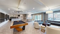 This generously sized single-floor amenity space offers an open-concept layout that seamlessly integrates a living area, dining room, and fully equipped kitchen. The interior design follows a contemporary aesthetic, characterized by coffered ceilings with recessed lighting, elegant pendant fixtures, and a consistent herringbone-patterned luxury vinyl flooring throughout.

The living area provides multiple seating arrangements, anchored by a wall-mounted flat-screen television and complemented by modern furnishings. A full-size pool table serves as a central recreational feature, positioned beneath a sleek ceiling fan. Textured, gray-toned accent walls add visual depth across the space.

The kitchen is outfitted with dark lower cabinetry, light wood upper cabinets, a black tile backsplash, stainless steel appliances, and an expansive white quartz island with bar seating illuminated by bold pendant lights. The dining area accommodates a large table for group gatherings and opens directly to an outdoor patio through full-length glass doors, allowing natural light to fill the space and providing convenient exterior access.