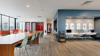 Located at 870 S Beechwood Ave in Fayetteville, Arkansas, this thoughtfully designed single-floor communal space offers a dynamic and versatile environment suited for both socializing and productivity. The open-plan layout is anchored by a large, warm-toned wood communal table surrounded by modern mint-green bar-height chairs, providing ample seating for gatherings or collaborative work sessions. A striking teal accent wall defines a dedicated workstation alcove, featuring a built-in desk fitted with desktop computers, patterned wallcovering, and framed wall art — creating a focused yet stylish work area. Recessed lighting throughout the bright white ceiling ensures consistent, well-distributed illumination across the space. Multicolored striped carpeting adds a contemporary, energetic touch underfoot. Floor-to-ceiling windows along one end of the space flood the interior with natural light and offer open views of the surrounding Fayetteville neighborhood. Bold pops of color — including red accent chairs and vibrant red double doors — punctuate the space with visual interest. Additional lounge seating and a small café-style area with a wet bar further enhance the space's multifunctional character, making it ideal for both casual relaxation and focused activity.