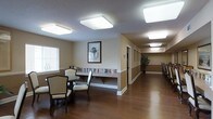 This single-story facility offers a thoughtfully designed common area that balances functionality with a warm, welcoming ambiance. The main gathering space features rich hardwood flooring that extends throughout, anchoring the room with a sense of warmth and continuity. Comfortable dining-style chairs and round tables provide seating for residents or guests, making the area well-suited for socializing or casual gatherings. A built-in shelf running along the lower portion of the wall houses an extensive collection of books and media, adding a library-like quality to the space. A framed piece of wall art depicting a palm tree adds a decorative focal point, complementing the neutral beige and tan wall tones. Large flush-mount ceiling light panels provide bright, even illumination throughout the space. Windows along one wall allow natural light to filter in through horizontal blinds, enhancing the room's openness. A wide, well-lit corridor extends from the main area, lined with additional framing and accented by a potted plant, suggesting further rooms and amenities beyond. The overall design is clean, accessible, and oriented toward comfort, making it an inviting environment for everyday use.