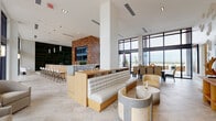 Located at 525 NE First Ave in Fort Lauderdale, FL, this contemporary amenity level is thoughtfully designed to offer residents a refined and multifunctional social environment. The space is anchored by soaring ceilings and expansive floor-to-ceiling windows that flood the interiors with natural light and frame views of the surrounding outdoor terrace and pool area.

The main lounge features warm wood cabinetry, custom banquette seating with geometric-patterned upholstery, and a mix of stylish lounge chairs and round accent tables, all set atop large-format travertine tile flooring. A dedicated bar area with counter seating and a dark stone accent wall adds a sophisticated hospitality feel, complemented by pendant lighting and a built-in media unit with open shelving.

A standout feature is the indoor putting green, flanked by a dramatic large-scale moss art wall with geometric copper detailing. A rich wood lattice partition elegantly separates zones while allowing light to pass through, and glass doors open directly to a furnished outdoor terrace with views toward the Fort Lauderdale skyline. A well-appointed kitchenette with stainless steel appliances and modern cabinetry rounds out the functional amenities. The overall ambiance is polished, airy, and inviting.