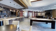 The amenity lounge at Woodmont Metro at Metuchen Station is a thoughtfully designed social space that blends modern sophistication with inviting comfort. The open-concept layout is anchored by large-format porcelain tile flooring arranged in a striking diagonal pattern, lending a sense of expansiveness to the room. A prominent billiards table serves as the centerpiece, making it an ideal gathering spot for residents. Adjacent to it, a sleek bar counter with high bar stools is illuminated by a series of stylish pendant lights suspended from a recessed ceiling tray, creating an intimate yet lively atmosphere. A wall-mounted flat-screen television is set against a rich, dark wood and brick tile accent wall, perfect for watching sports or entertainment. The space features bold burgundy accent walls that contrast elegantly with the crisp white ceiling and contemporary light fixtures. A semi-private dining area at the rear, enclosed with glass-paneled doors, offers a more intimate setting for gatherings or events. Additional pendant lighting throughout ensures the entire space is well-lit and welcoming. The combination of premium finishes, layered lighting, and versatile seating arrangements makes this lounge a standout amenity within the community.