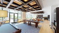 This thoughtfully designed single-floor amenity space offers a diverse range of gathering areas suited for both recreation and relaxation. The centerpiece game room impresses with a striking coffered ceiling accented by warm inset lighting, hardwood flooring, and large green-framed windows that flood the space with natural light. A full-size billiards table and shuffleboard table anchor the room, while a comfortable seating area with lounge chairs provides a casual retreat nearby.

The fully equipped kitchen features dark cabinetry, mosaic tile backsplashes, dark granite countertops, stainless steel appliances, and a breakfast bar with seating. Adjacent to the kitchen, a formal dining area accommodates a large dark wood table beneath a pendant light, with garden views through oversized windows.

A dedicated lounge area offers a built-in bench seating wall, while a separate sitting room showcases a floor-to-ceiling mosaic tile fireplace surround with a mounted TV, brick-patterned flooring, and curated artwork. Additional nook seating and art displays enhance the communal feel throughout.

Outside, a beautifully landscaped courtyard features herringbone brick paving, white pergolas, lush planted beds with flowering shrubs, and benches, creating a serene outdoor retreat.