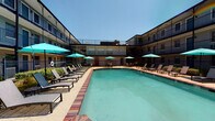 This amenity space features two distinct areas designed for recreation and fitness. The outdoor pool area is a welcoming retreat centered around a generously sized swimming pool with clear turquoise water, framed by a classic brick-tiled surround. Rows of sleek lounge chairs line both sides of the pool, complemented by multiple teal patio umbrellas providing ample shade. The courtyard setting is enclosed by a multi-story building with open walkways, lending a resort-like atmosphere under open skies. The fitness center is a comprehensively equipped space featuring herringbone-patterned carpet flooring and abundant natural light streaming through multiple large windows with views of the surrounding greenery. The gym is furnished with a full range of cardio equipment, including treadmills and a rowing machine, alongside an extensive strength training section featuring a power rack, cable machine, a complete dumbbell rack, medicine ball storage, battle ropes, and a Precor stationary bike. Large wall-to-wall mirrors expand the sense of space, while ceiling fans contribute to a comfortable workout environment. Recessed lighting and high ceilings further enhance the open, airy feel of the fitness area.