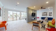 Located at 5010 W Broad St NE in Sugar Hill, Georgia, this thoughtfully designed single-floor community lounge offers an inviting and vibrant gathering space. The herringbone-patterned brick flooring flows seamlessly throughout, anchoring the eclectic mid-century modern aesthetic. A dramatic floor-to-ceiling tiled accent wall serves as the room's focal point, housing a sleek linear fireplace below and a mounted flat-screen TV above, accented by a decorative sunburst wall sconce. Recessed lighting and a stylish ceiling fan maintain a bright, comfortable atmosphere throughout. The lounge area is furnished with colorful, retro-inspired seating — including a chartreuse sofa with bold orange accent pillows — arranged around an industrial-style coffee table. Striking navy geometric lattice panels serve as eye-catching wall art, adding depth and design interest. Built-in open shelving with a wet bar area provides additional functionality. On the opposite end, floor-to-ceiling glass doors and expansive windows flood the space with natural light and offer direct views of and access to the outdoor pool deck and surrounding community, creating a seamless indoor-outdoor connection ideal for social gatherings and community events.