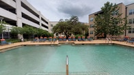 This outdoor pool area serves as a central amenity space, thoughtfully designed for relaxation and recreation. The generously sized swimming pool features clear turquoise water and concrete coping, with easy entry provided by built-in handrails. An attached spa sits adjacent to the main pool, offering a dedicated space for hydrotherapy. The expansive sundeck surrounding the pool is finished in a light-toned concrete surface and is well-equipped with a large collection of blue lounge chairs accented with orange cushions, arranged along the pool's edge to maximize sun exposure and poolside comfort. Mature trees are interspersed throughout the courtyard, providing natural shade and greenery that softens the hardscape. The pool area is enclosed by multi-story residential buildings, creating a sheltered, courtyard-like atmosphere. Access to the pool is through a dedicated entry point framed by dark wood-trimmed doors set into a stucco exterior wall, with posted pool rules nearby. Large glass windows and sliding doors from the adjacent pool house or amenity room offer direct sightlines to the pool deck, enhancing the sense of openness. The overall setting balances functional outdoor amenities with an inviting, park-like ambiance.