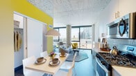 Located at 160 N Elizabeth Street in Chicago's vibrant West Loop neighborhood, this contemporary residence showcases a thoughtfully designed open-concept layout that seamlessly connects the kitchen, dining, and living areas. Exposed concrete ceilings lend an industrial character throughout, complemented by wide-plank hardwood flooring and a bold yellow accent wall that adds a striking pop of color.

The kitchen is well-appointed with stainless steel appliances — including a gas range, microwave, dishwasher, and refrigerator — paired with crisp white upper cabinetry, gray lower cabinets, and a classic subway tile backsplash. Track lighting illuminates the workspace efficiently. The adjacent dining area flows naturally into the living room, where floor-to-ceiling windows and a concrete structural column frame expansive, elevated views of the Chicago skyline and surrounding West Loop streetscape.

The bedroom continues the floor-to-ceiling window treatment, flooding the space with abundant natural light and offering sweeping city panoramas. The bathroom is finished with a gray vanity topped with a white countertop, an illuminated backlit mirror, and a glass-enclosed walk-in shower featuring clean white subway tile. Together, these elements create a refined, modern urban living experience defined by light, views, and contemporary design.