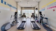 This single-floor fitness center is designed to inspire and support a well-rounded workout routine. The space is bright and open, characterized by high ceilings, light wood-style flooring, and oversized glass garage doors that span an entire wall, bringing in abundant natural light and offering views of the surrounding outdoor area, including a glimpse of a pool beyond.

The cardio section is generously equipped with multiple treadmills — including Precor models — along with elliptical trainers and a stationary bike, each machine offering ample space for comfortable use. A dedicated strength and functional training area features a cable machine, a weight bench, a spin bike, a rowing machine, and a stair climber, complemented by a full-length wall mirror that enhances the sense of depth throughout the room. Stability balls and medicine ball racks round out the functional fitness options.

Ceiling fans and recessed lighting ensure a comfortable environment at any time of day. Mounted televisions provide entertainment during workouts. The walls are adorned with bold, colorful motivational typography — words like "Dream," "Energize," "Determination," and "Fitness" — adding an energetic and uplifting ambiance. Overall, the space combines practicality with a dynamic, encouraging atmosphere suited for all fitness levels.