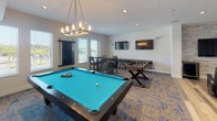 Located at 9511 Wes Kearney Way in Riverview, Florida, this single-floor community game and recreation room is thoughtfully designed to provide an engaging, social environment for all who use it. The space seamlessly blends two distinct flooring zones — warm, wide-plank hardwood and a stylish patterned carpet — that help define separate activity areas within the open layout.

At the heart of the carpeted zone sits a full-size pool table with a vibrant teal felt surface, illuminated by an elegant rectangular chandelier overhead. Nearby, a foosball table and wall-mounted flat-screen televisions add to the entertainment options, while a high-top dining table with chairs provides a casual spot to gather. A shuffleboard table is also present, offering yet another classic game option.

Transitioning to the hardwood section, plush leather recliner chairs and a rustic industrial dining table create a comfortable lounge and viewing area. This section is further enhanced by a classic arcade cabinet and a pinball machine, lending a nostalgic, fun atmosphere to the space. Recessed lighting throughout keeps the room bright and welcoming, complemented by natural light streaming in through multiple windows. The clean white walls serve as a neutral backdrop, accented by curated artwork. Additional rooms, visible through interior glass-panel doors, suggest access to further amenities beyond this recreation space.