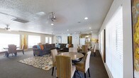 This single-floor space is designed as an open-plan gathering area that seamlessly integrates multiple functional zones. The living area is anchored by a large, plush sectional sofa in a warm charcoal tone, accented with teal and orange throw pillows, and positioned atop a bold, multicolored circular-patterned area rug. Ceiling fans with integrated lighting are evenly distributed across the textured ceiling, complemented by recessed lighting that ensures the space is well-lit throughout. Multiple windows along the walls invite natural light, contributing to the bright and airy atmosphere. The dining area features two distinct seating arrangements: a round wooden table with woven rattan-style chairs, and a larger rectangular table with matching seating, providing ample space for group gatherings. A partial view into the kitchenette reveals a clean, functional setup with white appliances and light cabinetry. The flooring throughout is a deep charcoal carpet that adds warmth and cohesion across the open layout. A wall-mounted clock and decorative artwork provide subtle personalized touches. Overall, the space conveys a casual yet organized communal ambiance, well-suited for socializing and shared activities.