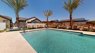 Located in north Phoenix, Arizona, this single-level outdoor community amenity area offers a resort-style experience designed for relaxation and recreation. At its centerpiece is a generously sized rectangular swimming pool with crystal-clear blue water, complemented by an adjacent attached spa. The pool surround is finished with light-toned pavers, providing ample space for sunbathing and circulation.

Lining both sides of the pool area are rows of cushioned chaise lounges, many shaded by vibrant blue market umbrellas, ideal for lounging on sunny Arizona days. Patches of manicured artificial turf add a pop of green against the desert landscape and serve as additional relaxation zones between the seating arrangements. Tall, mature palm trees are strategically positioned throughout the space, offering natural shade and lending the area a tropical, resort-like ambiance.

A pool house structure anchors one end of the space, featuring large windows, warm stucco exterior walls, and covered overhangs with exterior wall-mounted light fixtures. The entire area is enclosed by a combination of white privacy walls and wrought iron fencing, ensuring security while maintaining open views of the surrounding residential neighborhood. The space is designed to maximize Arizona's abundant sunshine while providing practical amenities for community use.