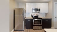 This well-appointed single-floor residence offers a thoughtfully designed open-concept layout that seamlessly connects the living and kitchen areas. The kitchen is a standout feature, boasting stainless steel appliances including a refrigerator, electric range, and over-the-range microwave. Crisp white upper cabinetry contrasts elegantly with dark lower cabinets, complemented by a subway tile backsplash and a spacious island with a double sink and white countertop surface — ideal for both meal preparation and casual dining. Light wood-tone flooring runs throughout the main living area, adding warmth and continuity to the space. The bedroom is generously sized, finished with plush neutral carpeting, a ceiling fan with light fixture, and a window that invites natural light. A dedicated hallway leads to a closet with built-in shelving and an in-unit laundry area, enhancing everyday convenience. The full bathroom is clean and functional, featuring large-format tile flooring, a dark espresso vanity with an integrated sink and mirror, and a combined shower-tub unit. Neutral warm-toned walls throughout the residence create a cohesive, inviting atmosphere suited for comfortable modern living.