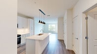 This thoughtfully designed 2-bedroom, 2-bathroom residence offers a functional and modern single-floor layout. The open-concept kitchen serves as the heart of the home, showcasing white shaker-style cabinetry extending to the ceiling, stainless steel appliances including a refrigerator with ice maker, gas range, and built-in microwave. A large center island with a white quartz countertop and undermount sink is illuminated by striking matte black pendant lights, creating a stylish focal point. Hardwood-style flooring flows seamlessly throughout the space, connecting the kitchen to a bright and open living area with large windows that invite abundant natural light and outdoor views. The spacious primary bedroom features its own walk-in closet and is flooded with natural light from dual windows overlooking the surrounding neighborhood. The en-suite primary bathroom is outfitted with a double vanity, backlit mirror, white cabinetry, and a separate shower. The second bathroom includes a full tub-shower combination with white tile surround. The second bedroom also benefits from large windows and generous floor space. A dedicated hallway provides smooth circulation between all rooms, completing this clean, contemporary residence.