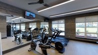 Located at 1-3 Mill River Lane in Rads let, NY, this well-appointed fitness center offers a clean, modern environment designed to support a comprehensive workout routine. The space is laid out on a single floor, maximizing accessibility and ease of use for all visitors. The room is equipped with a variety of cardio machines, including treadmills, ellipticals, and stationary bikes, all arranged to allow ample space between each station. Full-length mirrored walls line one side of the room, enhancing the sense of openness while allowing users to monitor their form during exercise. The light wood-toned flooring adds warmth to the space and contrasts tastefully with the dark, textured accent wall panels that lend a contemporary aesthetic. Overhead, sleek linear LED lighting and a ceiling fan ensure the space remains well-lit and well-ventilated. Large windows along the exterior wall invite an abundance of natural light and provide views to the outdoors, creating an energizing atmosphere. A wall-mounted flat-screen television is positioned for easy viewing during workouts, adding an entertainment option for users. Overall, the space strikes a balance between functional fitness amenities and modern interior design.
