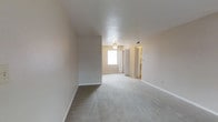This single-floor residential space offers a clean and straightforward layout designed for comfortable everyday living. The interior is finished with wall-to-wall carpet in a neutral gray tone that extends throughout the main living areas, contributing to a warm and quiet atmosphere underfoot. Walls are painted in soft, light beige tones that complement the bright, airy feel of the space.

The open living area is generously proportioned, allowing for flexible furniture arrangements. A ceiling fan is centrally positioned overhead, providing both air circulation and a subtle design element. Natural light filters in through a window at the far end of the room, illuminating the space and enhancing its open character.

A partial dividing wall adds a degree of architectural definition between areas without fully enclosing the space, maintaining an open flow. An adjacent doorway, trimmed in white, leads to a connecting room or hallway, with a glimpse of additional space beyond — including what appears to be a closet with bifold doors and a warmly lit adjacent area. HVAC vents are integrated into the ceiling, ensuring climate control throughout. White baseboards frame the floors cleanly, adding a crisp finish to the overall interior. The space presents a move-in-ready, functional environment with a calm, neutral aesthetic.