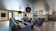 This thoughtfully designed single-floor amenity space offers a seamless blend of style and functionality across several distinct yet interconnected zones. The main lounge area commands immediate attention with its dramatic dark metallic ceiling, anchored by striking oversized spherical pendant lights that cast a warm, ambient glow over plush sectional sofas and leather lounge chairs arranged on richly patterned area rugs. Floor-to-ceiling sliding glass doors line the exterior wall, flooding the space with natural light and providing direct views and access to a landscaped outdoor courtyard. A dedicated seating alcove near the entry features tufted upholstered sofas alongside warm wood accent columns, offering a more intimate gathering space. The kitchen and bar area is appointed with a generous white stone island, upholstered counter seating, matte black cabinetry, a textured tile backsplash, and open shelving adorned with lush greenery — all illuminated by a trio of bold dome pendant lights. An adjacent lounge section showcases light hardwood flooring, curated wall art, tall potted plants, and a mix of blue and tan upholstered seating, creating a relaxed yet refined atmosphere throughout. The exterior model reveals a multi-story building wrapped around a central landscaped courtyard.