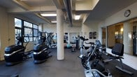 This fitness center is a thoughtfully designed, single-floor facility offering a comprehensive workout environment. The space features an open layout supported by prominent cylindrical columns, with exposed industrial-style ductwork running across the ceiling, lending the room a modern, utilitarian aesthetic. Recessed skylights and large windows along the perimeter flood the floor with natural light, creating a bright and energizing atmosphere.

The gym is well-stocked with a diverse selection of equipment to accommodate a full range of fitness routines. Cardio machines, including stationary bikes, an elliptical, and a stair climber, are positioned throughout the space for easy access. A dedicated free weights area features a full rack of dumbbells, while stability balls and functional training accessories are available for varied workout options. Strength and resistance machines are arranged along the walls, maximizing floor space efficiency.

A wall of mirrors on one end enhances the sense of openness and supports form-checking during exercise. The dark rubberized flooring provides both durability and safety underfoot. An informational bulletin board near the entrance offers posted guidelines and resources. Overall, this fitness center delivers a clean, functional, and well-lit environment suitable for a broad range of fitness levels and training goals.