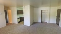 This single-floor residential space presents a straightforward and functional layout designed for comfortable everyday living. The main living area features neutral-toned carpeting that adds warmth underfoot, while the plain white walls provide a clean, bright backdrop throughout. The open layout creates a sense of spaciousness, with the living area flowing naturally toward the kitchen. The kitchen is partially separated by a half-wall partition, offering a degree of definition between spaces while maintaining an open feel. It is equipped with dark brown cabinetry that provides ample storage, a dishwasher, and a sink set beneath the cabinetry, all finished with tile flooring that contrasts with the carpeted living area. A floor vent is visible, indicating a built-in heating and cooling system. On the opposite side of the living area, a closet with a built-in shelf and hanging rod offers practical storage solutions. The overall ambiance of the space is simple and utilitarian, with neutral tones and minimal detailing throughout, making it a versatile canvas well-suited for personalization.