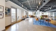 Located at 120 Border St in Boston's East Boston neighborhood, this expansive single-floor commercial space offers a striking industrial-chic aesthetic. The open-plan layout is defined by polished concrete floors that reflect natural light throughout, while the exposed ceiling reveals structural beams, ductwork, and conduit painted in dark tones — a hallmark of modern loft-style design. Large windows line the perimeter, flooding the interior with abundant natural light and framing views of the surrounding waterfront and Boston skyline, including a notable glimpse of the Tobin Bridge. The space is divided into distinct functional zones, including lounge areas furnished with contemporary sofas and accent chairs, casual dining and bar-height seating along a wood-topped counter, and open collaborative areas with tables and modern chairs. Curated artwork adorns the white walls throughout, lending a gallery-like quality to the environment. A private outdoor terrace accessed through glass doors offers additional waterfront views. Private rooms with grey doors provide enclosed spaces off the main floor. The overall ambiance is dynamic and versatile, blending urban industrial character with polished, thoughtfully designed interiors — well-suited for collaborative work, events, or creative endeavors.