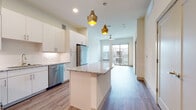 This well-designed single-floor residence combines contemporary style with functional living. The open-concept kitchen is a standout feature, showcasing crisp white cabinetry, a full suite of stainless steel appliances including a refrigerator, range, microwave, and dishwasher, and a granite-topped island with pendant lighting in brushed gold tones. Hardwood-style flooring runs throughout the main living areas, contributing to a clean and cohesive aesthetic. The kitchen flows seamlessly into the living area, which is capped by a ceiling fan and opens onto a private balcony through a sliding glass door, allowing for natural light and outdoor access. The bedrooms are carpeted for comfort and receive ample natural light through large windows fitted with blinds. One bedroom features direct access to an exterior door, adding versatility. The bathroom is finished with white subway tile surrounding the tub/shower combination, wood-style flooring, a granite vanity, and white cabinetry for storage. A staircase visible in the model suggests additional access points within the building. The overall ambiance is modern, light, and airy, with a neutral palette throughout that offers a welcoming and move-in-ready environment.