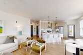 This well-designed single-story home offers a thoughtful layout across two bedrooms, one bathroom, and a seamlessly connected living, dining, and kitchen area. The open-concept main living space is bright and airy, featuring light hardwood-style flooring, white walls, and a ceiling fan that enhances the comfortable ambiance. The kitchen is a standout feature, showcasing light wood cabinetry, a white tile backsplash, stainless steel appliances including a range with a sleek hood vent, and a central island with bar seating illuminated by stylish brass pendant lights. Adjacent to the kitchen, the dining area accommodates a round marble-top table beneath a modern multi-bulb chandelier. The living room benefits from natural light through windows offering outdoor views. The primary bedroom is a comfortable retreat with plush carpeting, a bright window, and clean white walls. The bathroom features light wood vanity cabinetry with white countertops and hardwood-style flooring. Two closets provide ample storage, and a hallway connects the main spaces efficiently. The attached garage includes laundry hookups, adding everyday convenience. Overall, the home presents a clean, modern aesthetic with cohesive finishes throughout.
