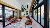 Located at 9840 Carroll Canyon Rd in San Diego's Scripps Ranch community, this contemporary amenity lounge is designed to impress. The single-floor space features soaring ceilings with warm wood paneling and recessed lighting, creating an airy, upscale atmosphere throughout. Light hardwood flooring runs the length of the lounge, complementing the clean, modern aesthetic.

The main lounge area is furnished with cognac leather armchairs and sleek black metal accent tables, arranged atop a textured area rug to create inviting conversation zones. Living green wall installations add a striking organic element, while bold striped wall detailing provides visual contrast. Floor-to-ceiling black-framed glass walls and doors flood the interior with natural light and offer sweeping views of the outdoor pool deck, complete with lounge chairs and palm trees beyond.

A dedicated dining and collaboration area features built-in booth seating with geometric-tiled walls, warm wood slatted dividers, and pendant lighting — ideal for casual meetings or private gatherings. A kitchenette with warm wood cabinetry, quartz countertops, and a stainless steel undermount sink provides functional convenience. An additional seating nook with floor-to-ceiling windows frames views of the landscaped surroundings and neighboring streetscape, filling the space with abundant Southern California sunlight.