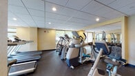 This fitness center offers a spacious, single-floor layout designed to accommodate a full range of workout needs. The room is finished with a drop tile ceiling fitted with recessed lighting, while warm yellow-toned walls create an energizing atmosphere throughout the space. Durable rubber flooring covers the entire workout area, providing both safety and comfort underfoot.

The cardio section is well-appointed with multiple treadmills, elliptical trainers, and a stair climber, all arranged to take advantage of the large floor-to-ceiling windows that line the perimeter walls. These expansive windows flood the space with natural light and offer views of the surrounding outdoor greenery, making workouts more enjoyable.

The strength training area features a variety of selectorized cable machines and weight equipment, catering to a full-body workout routine. A wall-mounted television is positioned near the strength equipment for added convenience. Mirrors are strategically placed to support proper form during exercises.

The facility also includes an adjacent restroom accessible via a glass-paneled door, with a wooden bench positioned in the entryway for added convenience. The overall layout is open and uncluttered, allowing for easy movement between equipment zones and ensuring a comfortable, focused exercise environment.