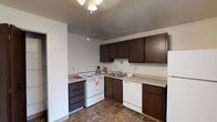 Located at 69 Jadwin Avenue in Richland, Washington, this single-floor unit offers a straightforward and functional layout designed for comfortable everyday living. The kitchen is well-equipped with dark wood cabinetry providing ample storage, light-toned countertops, and white appliances including an electric range, dishwasher, and refrigerator. Stone-patterned tile flooring runs throughout the kitchen and entry areas, offering durability and easy maintenance. The living area features wall-to-wall carpeting and notably elevated ceilings that lend a sense of openness to the space. Large windows allow natural light to filter in, creating a bright and airy atmosphere. The layout transitions smoothly from the living area through a connecting hallway toward the bedroom and bathroom. The bedroom is carpeted, with a window that brings in additional natural light and an exterior view of the surrounding neighborhood. Baseboard heating units are present in multiple rooms, ensuring efficient climate control throughout. The full bathroom includes a tub/shower combination, toilet, and vanity with matching dark wood cabinetry. Additional storage is available through a dedicated closet space with built-in shelving and a hanging rod, adding practical utility to the overall floor plan.