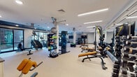 Located at 510 E Pettigrew St in Durham, NC, this modern fitness center offers a comprehensive and thoughtfully designed workout facility across a single floor. The space is divided into distinct functional zones, each catering to different training needs. The main strength training area features a full rack of dumbbells, multiple adjustable benches, a power rack with a cable attachment system, and wall-to-wall mirrors that enhance the sense of space and allow users to monitor their form. Linear LED lighting runs across the ceiling, providing bright, even illumination throughout. A dedicated cardio zone houses a row of treadmills, elliptical machines, and a stair climber, accompanied by a wall-mounted flat-screen TV and ceiling fans for comfort. An additional area near large sliding glass doors provides natural light and outdoor views, and is equipped with a rowing machine and stair climber, creating an inviting environment for lower-intensity or warm-up training. Textured accent walls and a dark-paneled ceiling in select areas add a contemporary design element that contrasts with the otherwise clean, neutral palette. The flooring throughout consists of durable, light-toned carpet suitable for a high-use athletic environment. The overall ambiance is professional, modern, and well-lit, offering a fully functional facility for a wide range of fitness routines.