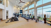 This expansive fitness center is thoughtfully designed to accommodate a full range of exercise disciplines across multiple zones. The main cardio area is defined by soaring double-height ceilings, large ceiling fans, and an impressive wall of floor-to-ceiling windows that flood the space with natural light and offer open views of the outdoor amenity area. A generous row of treadmills and elliptical machines lines this bright, airy section, complemented by rowing machines and additional cardio equipment.

Adjacent to the cardio floor, a dedicated strength training zone features free weights including multiple dumbbell racks, a power rack, cable machines, and a multi-function strength station. A textured accent wall and wall-mounted television add visual interest to this area, which is finished with low-pile carpet tiles throughout.

A separate studio room provides a more intimate setting for focused workouts, with wood-look flooring, a wall-mounted television, spin bikes, mirrored walls with glass partitions, and built-in shelving stocked with exercise balls. A water bottle filling station is conveniently located at the transition between zones. The facility is finished in a clean, contemporary palette of whites and warm neutrals, creating a bright and motivating environment throughout.