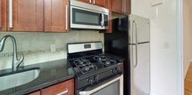 This single-floor residence offers a thoughtfully designed layout that balances comfort and functionality. The kitchen is outfitted with stainless steel appliances, including a gas range, over-the-range microwave, and refrigerator, complemented by rich dark-stained cabinetry, dark granite countertops, and a neutral tile backsplash that together create a polished, practical cooking space. The living area features warm-toned hardwood flooring that extends throughout the residence, crisp white walls with clean crown molding, and a welcoming entryway with a solid front door. Multiple bedrooms offer generous proportions, each bathed in natural light through double-hung windows that frame lush greenery outside. The bedrooms are finished with the same gleaming hardwood floors, and at least one includes a dedicated closet, providing ample storage. Radiator heating units are present in several rooms, adding a classic character to the space. The bathroom is elegantly appointed with floor-to-ceiling neutral ceramic tile, a soaking bathtub with shower, a dark-stained vanity with an undermount sink, and a frosted window that allows natural light while maintaining privacy. Throughout the residence, a clean, neutral palette and quality finishes create a bright and inviting atmosphere.