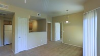 This single-story space features an open-concept layout that connects the main living area to the kitchen through a convenient pass-through counter, providing both visual connectivity and functional separation. The living area is finished with wall-to-wall carpeting in a neutral beige tone, complementing the warm, earthy paint on the walls throughout. A pendant light fixture with a classic dome shade hangs prominently, serving as a focal point while providing ambient lighting to the space. The kitchen, visible through the pass-through opening, features recessed track lighting and wood-toned cabinetry, offering a practical and efficient workspace. The ceiling is finished with a clean, flat surface accented by subtle crown-like detailing, adding a touch of architectural refinement. A centrally located doorway leads to additional rooms beyond, suggesting a functional floor plan with clear room separation. Ample wall space and multiple electrical outlets are distributed throughout, supporting versatile furniture arrangements and everyday use. The overall ambiance is clean, neutral, and move-in ready, with a cohesive color palette that lends a calm, understated character to the interior.