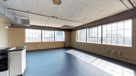 This single-floor industrial space offers a compelling blend of raw character and open-plan functionality. The interior is defined by its painted brick walls, which lend a textured, urban aesthetic throughout. Original wood-plank ceilings, finished in white, add warmth and historical depth overhead, while exposed steel beams underscore the building's structural heritage and industrial roots.

Large, multi-pane steel-framed windows line two walls of the space, flooding the interior with generous natural light. The windows also frame views of the surrounding cityscape, providing a dynamic urban backdrop. The interplay of sunlight through the grid-patterned windows casts striking geometric shadows across the smooth, blue-grey concrete floor, adding visual interest to the open expanse.

The layout is entirely open, making the space highly versatile and adaptable for a variety of uses, from creative studio or gallery applications to office or event configurations. Exposed utility conduits run along the walls, consistent with the industrial character of the space. The overall ambiance is one of understated, loft-style sophistication — raw and functional, yet full of character and flooded with light.
