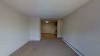 This single-floor unit offers a straightforward and functional layout designed for practical, everyday living. The main living area is carpeted throughout, providing a soft underfoot feel, and is defined by clean white walls and a flat white ceiling that lend a simple, neutral aesthetic. The open layout allows the living space to flow naturally toward the kitchen and entryway area, creating a sense of connectivity between the rooms.

The kitchen is positioned at the rear of the unit and is equipped with essential appliances, including a range and a refrigerator. A door adjacent to the kitchen serves as the primary entry point into the unit. Overhead lighting in the kitchen area provides functional illumination for meal preparation.

A baseboard heating unit runs along one wall of the living area, offering an efficient and unobtrusive heating solution throughout the space. The walls throughout are smooth and painted in a consistent neutral white, keeping the interior bright and easy to customize.

Overall, the unit presents a clean, no-frills interior that is move-in ready. Its simple design and open flow make it a practical choice for those seeking an uncomplicated, functional living environment on a single level.
