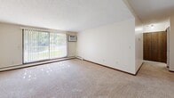 Located in Roseville, MN, this one-bedroom, one-bathroom unit offers a straightforward single-floor layout designed for comfortable everyday living. The living area is generously proportioned, featuring wall-to-wall carpeting, neutral cream-toned walls, and a large sliding glass door that floods the room with natural light while offering views of the surrounding greenery. A wall-mounted air conditioning unit and baseboard heating provide year-round climate comfort. The spacious bedroom continues the carpeted flooring and clean aesthetic, with a wide window fitted with blinds that allow for adjustable natural light. Baseboard heating runs along the perimeter, and the room offers ample space for furnishings. A connecting hallway links the main living spaces and provides access to cabinetry for additional storage. The bathroom features a full tub-and-shower combination with clean white square tile surround, maintaining a simple, functional design. Throughout the unit, dark wood trim accents provide contrast against the light walls and ceilings, lending a cohesive, classic feel. The overall layout is efficient and well-organized, making practical use of the available space while maintaining a bright, livable atmosphere.