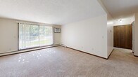 Located in Roseville, MN, this one-bedroom, one-bathroom unit offers a straightforward single-floor layout designed for comfortable everyday living. The living area is generously proportioned, featuring wall-to-wall carpeting, neutral cream-toned walls, and a large sliding glass door that floods the room with natural light while offering views of the surrounding greenery. A wall-mounted air conditioning unit and baseboard heating provide year-round climate comfort. The spacious bedroom continues the carpeted flooring and clean aesthetic, with a wide window fitted with blinds that allow for adjustable natural light. Baseboard heating runs along the perimeter, and the room offers ample space for furnishings. A connecting hallway links the main living spaces and provides access to cabinetry for additional storage. The bathroom features a full tub-and-shower combination with clean white square tile surround, maintaining a simple, functional design. Throughout the unit, dark wood trim accents provide contrast against the light walls and ceilings, lending a cohesive, classic feel. The overall layout is efficient and well-organized, making practical use of the available space while maintaining a bright, livable atmosphere.