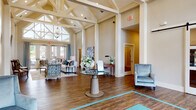 Located at 100 Legends Dr in Canton, GA, this single-story clubhouse and leasing center makes a striking first impression with its dramatic cathedral ceilings and intricately detailed exposed white beam truss system that defines the architectural character of the space. Rich hardwood-style flooring runs throughout, providing a warm contrast to the neutral, creamy wall tones that lend an air of sophistication and openness.

The grand main hall serves as the centerpiece of the space, anchored by a round glass-top entry table adorned with fresh floral arrangements. Soaring clerestory windows at the entrance flood the interior with generous natural light, while a statement chandelier adds an elegant touch above the main seating area. Comfortable lounge seating, featuring patterned accent chairs and a tufted sofa in coordinating teal and neutral tones, creates an inviting reception atmosphere.

A dedicated conference or consultation room offers a more intimate setting, appointed with a large oval table, upholstered seating, decorative drapery, and a classic arched mirror above a console. Decorative wall medallions and wall-mounted flat-screen televisions further enhance the common areas. French doors with floor-to-ceiling glass panels open to an outdoor courtyard, seamlessly connecting the interior to the surrounding community. The overall ambiance is polished, welcoming, and thoughtfully designed.