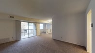 This single-floor residence offers an open, flowing layout that seamlessly connects its living areas. The main living space is anchored by a sliding glass door fitted with vertical blinds, opening onto a private balcony with a railed surround that welcomes natural light and provides outdoor views of surrounding greenery. The carpeted flooring runs throughout the interior, adding warmth and comfort underfoot.

The space transitions naturally into a secondary area, where a structural column and a low dividing wall with integrated built-in shelving define the boundary between zones without fully closing off the room. This built-in unit provides practical display and storage opportunities while maintaining the open, airy feel of the floor plan. A window in the adjoining area further contributes to the natural light that fills the space.

The interior finishes are clean and neutral, with white walls and light-toned ceilings that enhance the sense of openness. Flush ceiling lighting provides ambient illumination throughout. The overall layout is practical and versatile, offering residents flexibility in how they arrange and use the connected living areas. The combination of natural light, outdoor access, and thoughtful built-in features makes this a functional and comfortable single-floor living environment.