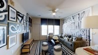 This single-floor living space showcases a thoughtfully designed interior with a strong monochromatic aesthetic. The living room serves as the focal point, anchored by a bold black-and-white graphic accent wall that adds dramatic visual interest. Large windows fitted with dark roller shades allow ample natural light to filter into the room, brightening the neutral-toned walls and light ceiling. A ceiling fan with an integrated light fixture is centrally positioned, providing both airflow and additional illumination.

The living area is furnished with a generously sized gray sectional sofa adorned with patterned throw pillows, complemented by a matching armchair that reinforces the cohesive design scheme. A geometric black-and-white area rug defines the seating zone, while two cylindrical black-and-white ottomans serve as functional accent pieces. A sleek rectangular coffee table anchors the arrangement, topped with a fresh floral centerpiece.

On the opposite wall, a flat-screen television is mounted on a low-profile media console with open storage. The surrounding wall features an eclectic gallery of framed black-and-white artwork, further unifying the room's sophisticated and curated monochromatic theme.