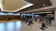 This single-floor spin studio is thoughtfully designed to provide an immersive and energizing workout environment. The room accommodates multiple stationary cycling bikes arranged in rows, offering ample space for group fitness classes. A full-length mirrored wall lines one side of the studio, visually expanding the space and allowing participants to monitor their form during sessions. The ceiling features a striking two-tone design, with a recessed tray detail accented by warm indirect lighting that contrasts with the darker surrounding panels, creating a layered and sophisticated look. Recessed downlights are evenly distributed throughout, ensuring the space is well-lit without being harsh. Dark wood-toned wall panels lend a refined, modern aesthetic, while a mounted flat-screen television serves as a focal point for on-screen workout guidance. Frosted or glazed windows along one wall allow for a degree of natural light while maintaining privacy. The light-toned flooring provides a clean, neutral base that complements the overall interior palette. The studio's design balances functionality with a polished ambiance suitable for a high-end fitness facility.