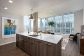 This contemporary urban residence offers a thoughtfully designed open-concept layout with high-end finishes throughout. The kitchen serves as the heart of the home, anchored by a large center island topped with striking white quartz countertops featuring subtle grey veining. Dark wood cabinetry pairs elegantly with stainless steel appliances, including a built-in microwave, range, and refrigerator, while a pendant light in brushed gold adds a refined touch. The kitchen flows seamlessly into the living area, where expansive floor-to-ceiling windows flood the space with natural light and frame sweeping views of the surrounding cityscape. An exposed concrete column adds an industrial architectural accent to the open living space. The primary bedroom continues the theme of abundant natural light, with large windows overlooking the urban skyline and plush carpeting underfoot. A second bedroom offers similarly generous light and city views. Two well-appointed bathrooms complete the layout — one featuring a soaking tub with marble-style surround, the other with a glass-enclosed walk-in shower, dark wood cabinetry, and wood-look flooring. Throughout, the residence strikes a refined balance between modern industrial design and warm, comfortable living.